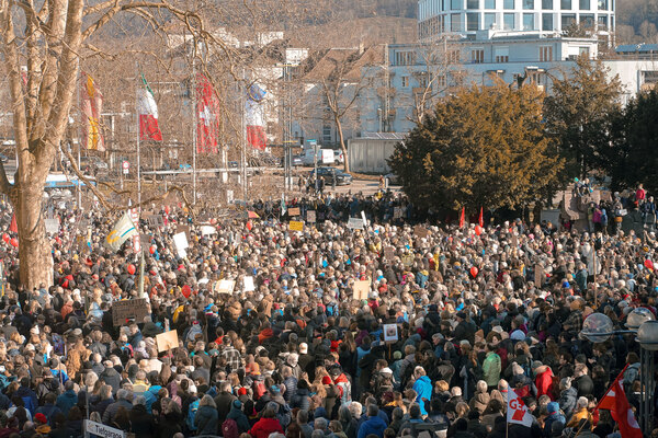Eine große Menschenmenge versammelt sich im Freien zu einem Protest. Die Menschen halten Schilder und Fahnen hoch, umgeben von Bäumen und städtischen Gebäuden im Hintergrund.